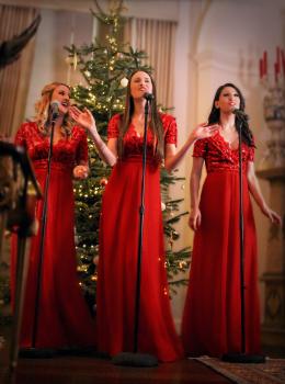 Three female singers in long red gowns in front of a christmas tree