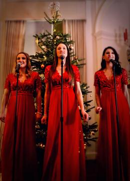 Three female singers in long red gowns in front of a christmas tree